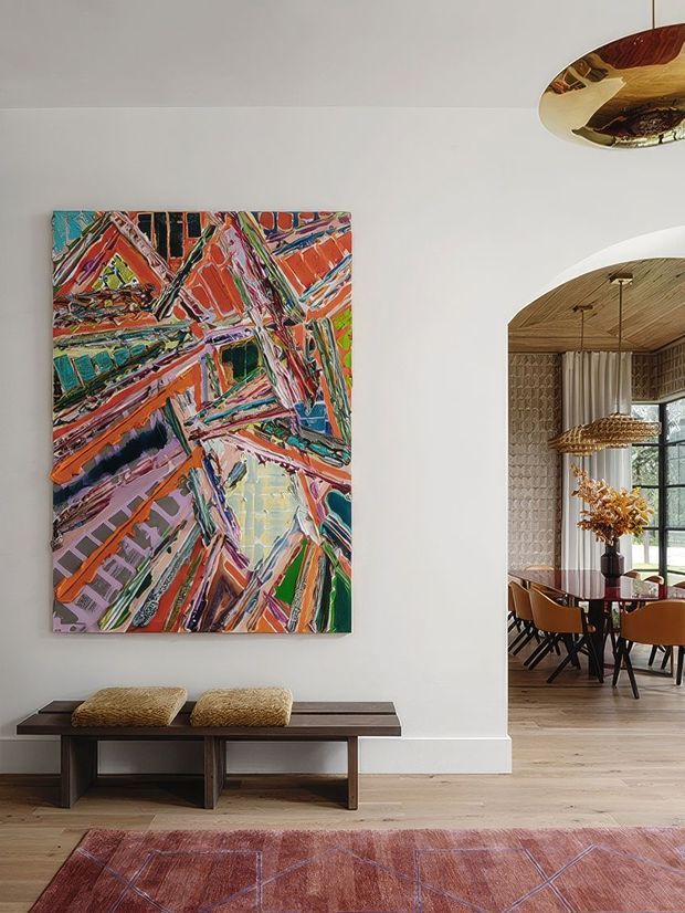 A refined interior view of a tall white-walled hallway or entry space, featuring a large vivid abstract painting above a dark timber bench with textured cushions. A pale timber floor and patterned rug add warmth. Through a wide arched opening, a dining room is visible with a timber ceiling, textured wall finish, pendant light, curtains, large window, and timber dining table with upholstered chairs. A sculptural ceiling light is partially visible in the foreground.