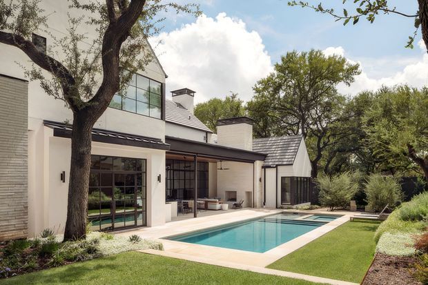 Exterior view of a refined contemporary residence surrounding a rectangular swimming pool and paved terrace. The home combines light rendered and brick-like wall surfaces, extensive dark-framed glazing, low-pitched metal roofs, and gabled volumes, set within a landscaped garden with mature trees and clipped lawn.