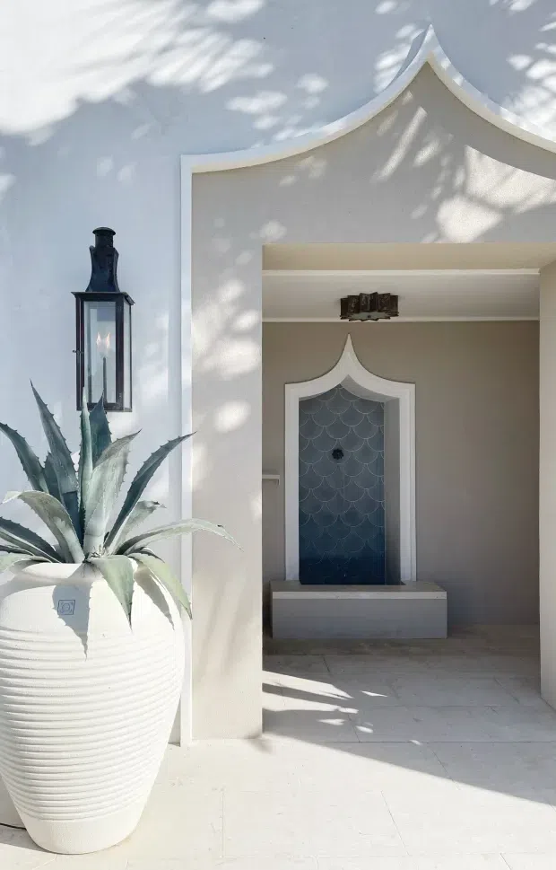 A bright exterior courtyard or entry alcove with white rendered walls and a distinctive curved, pointed decorative parapet. A large ribbed white planter with an agave stands beside a traditional dark metal wall lantern. The recessed alcove contains a small decorative water feature or fountain faced with blue scalloped tiles and framed by ornate pointed trim. Pale stone or tile paving and strong dappled sunlight create a refined, Mediterranean-influenced atmosphere.