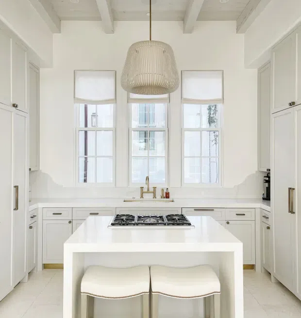 Bright, symmetrical white kitchen with a central island featuring an integrated gas cooktop and two upholstered stools. The room has extensive white cabinetry, pale stone or quartz work surfaces, three sash-style windows with roller shades, brass-toned tapware and hardware, and a large woven pendant light. The ceiling has exposed painted beams, creating a refined traditional architectural character with light contemporary styling.