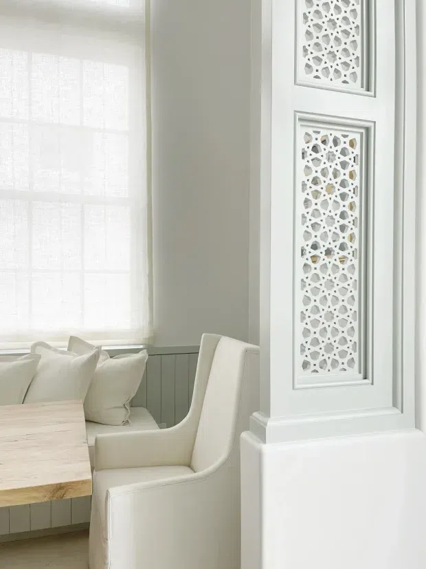 Bright, refined interior corner featuring a pale wood dining table, upholstered cream armchairs, a window seat with cushions, sheer window coverings, light wall panelling, and an ornate white geometric decorative grille inset into a column or wall panel. The palette is predominantly soft white and off-white with subtle timber accents.