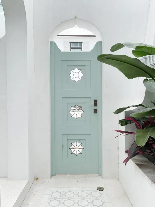 A bright, white-rendered entryway featuring a pale sage-green paneled front door with three decorative geometric star-shaped glazed inserts and dark hardware. The door is set within a pointed, scalloped arch opening, with a small transom window above. White tiled flooring includes a repeating geometric star pattern near the threshold, and a raised planter filled with tropical foliage borders the right side.