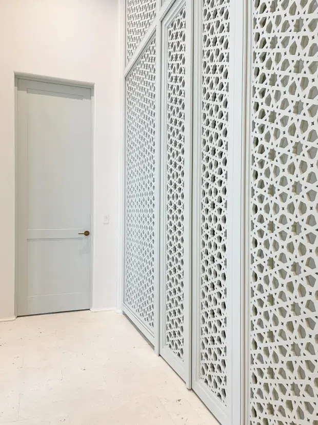 A bright, minimalist interior hallway featuring a pale grey-white paneled door with a small brass handle and a long decorative white geometric lattice screen in framed panels along the right wall. The space has white walls, light stone or tiled flooring, and abundant soft natural light. The latticework is the dominant architectural feature, providing privacy and visual texture.