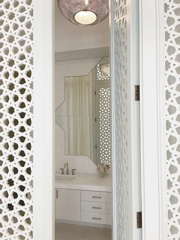 A bright, refined bathroom viewed through partially open white patterned privacy doors. The room features a floating or streamlined white vanity with drawers, a pale benchtop, brass-toned tapware, a large geometric-edged mirror, and a decorative perforated pendant light. White geometric screens and mirrored patterned details create an ornate, cohesive feature treatment.