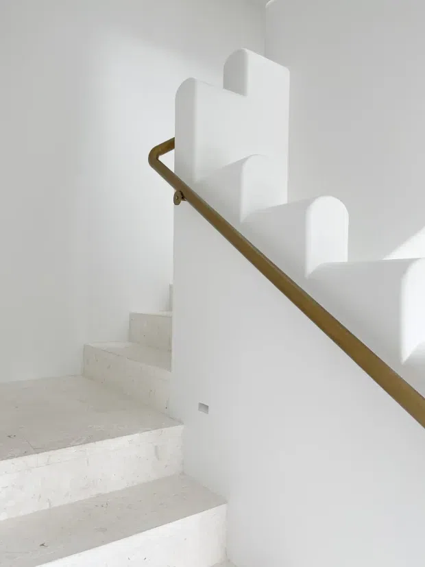 Minimalist interior stairway with broad white stone or concrete steps, white plaster walls, and a simple warm brass or bronze handrail. The stair balustrade features sculptural rounded vertical forms, creating a clean, softly geometric composition with bright natural light and very limited decoration.