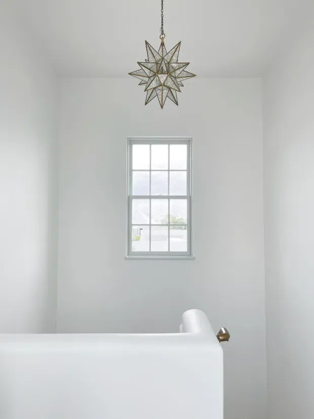Bright, minimalist bathroom with a freestanding white bathtub in the foreground, a white-painted wall, a single divided-pane window, and a decorative star-shaped pendant light suspended above. A small brass-toned bathtub fixture adds a subtle accent.