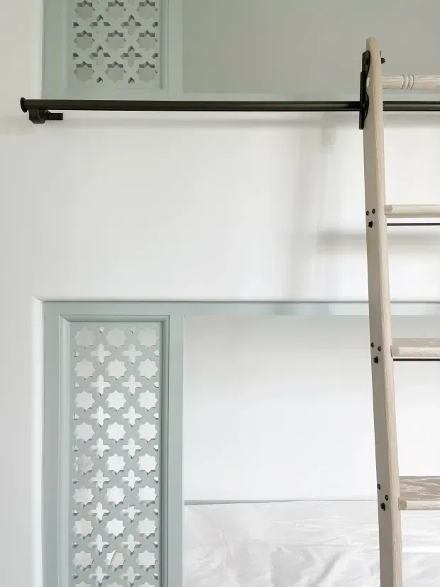 Close-up interior detail of a light, built-in bunk or loft bed structure. The image shows white painted wall panelling with decorative geometric fretwork inserts, dark metal horizontal rails, and a pale timber ladder leading to upper sleeping platforms. The styling is bright and refined with subtle coastal or traditional detailing.