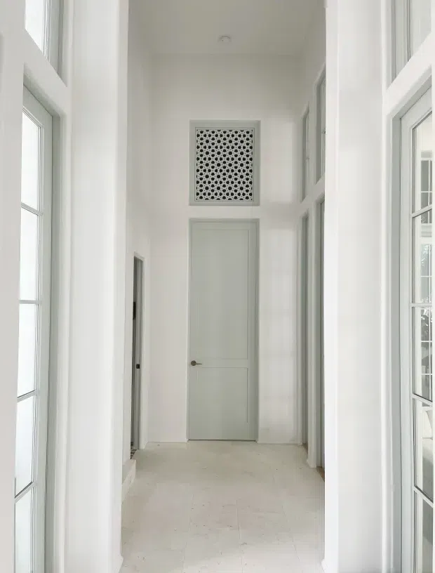 Bright, narrow residential hallway with white walls, tall white-framed glazed openings and doors on both sides, a pale tiled floor, and a simple closed interior door at the end. A decorative perforated grille is positioned above the end door, creating a clean, airy, highly symmetrical transitional space.