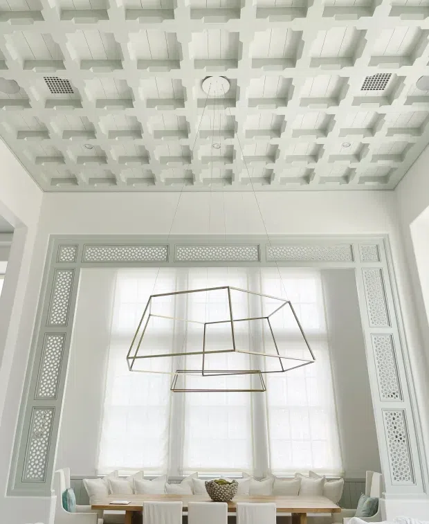 Bright, refined interior view dominated by a tall window framed with ornate geometric perforated decorative panels. A large sculptural geometric pendant hangs over a dining table with upholstered chairs and pale cushions. The ceiling features an extensive white coffered or three-dimensional geometric grid with recessed lighting and vents. The palette is predominantly white and off-white with subtle pale timber and metallic accents.
