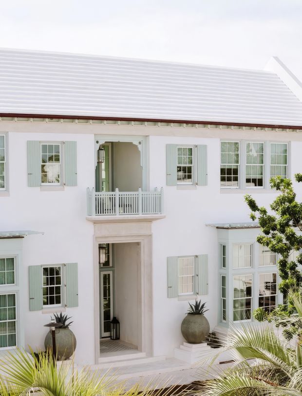 Bright exterior view of a refined two-storey white residence with a pale metal pitched roof, symmetrical multi-pane windows, pale green shutters, a recessed central entry, and a small white balustraded balcony above the doorway. Tropical planting and palms frame the foreground, giving the facade a coastal, resort-like character.