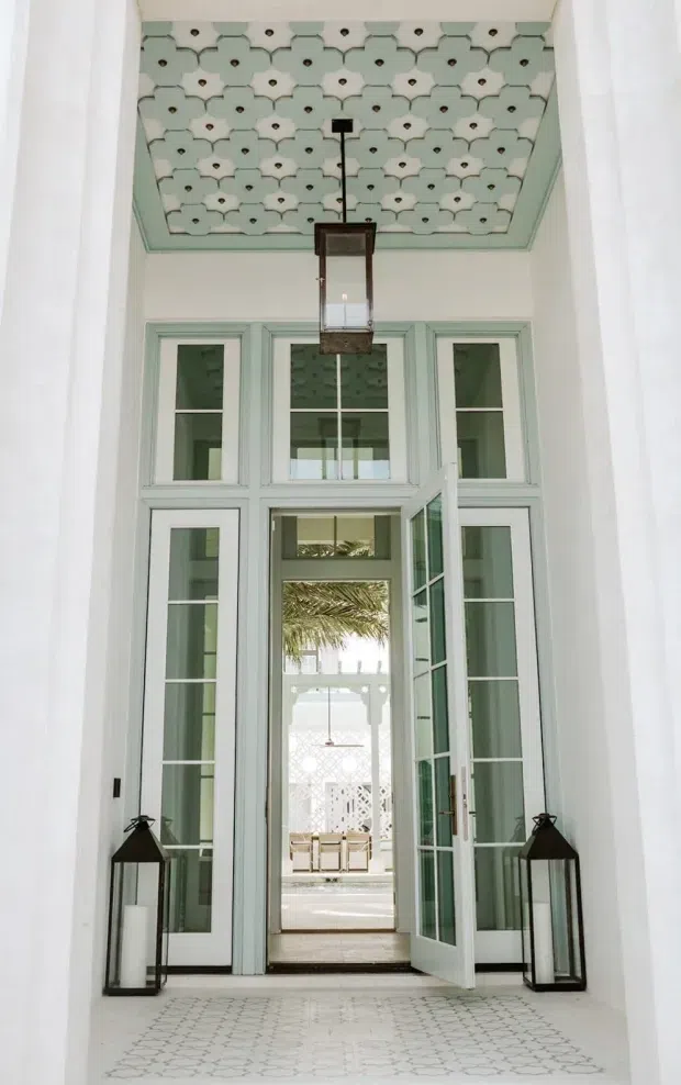 A tall, bright entry vestibule with pale mint-green French doors and sidelights, opening toward a sunlit courtyard. The space features a highly decorative patterned pressed-tin or plaster ceiling with matching mint trim, a suspended rectangular lantern, white walls, patterned light tile flooring, and two dark floor lanterns. The architecture has strong historic decorative detailing and a refined Mediterranean or Moroccan-inspired character.