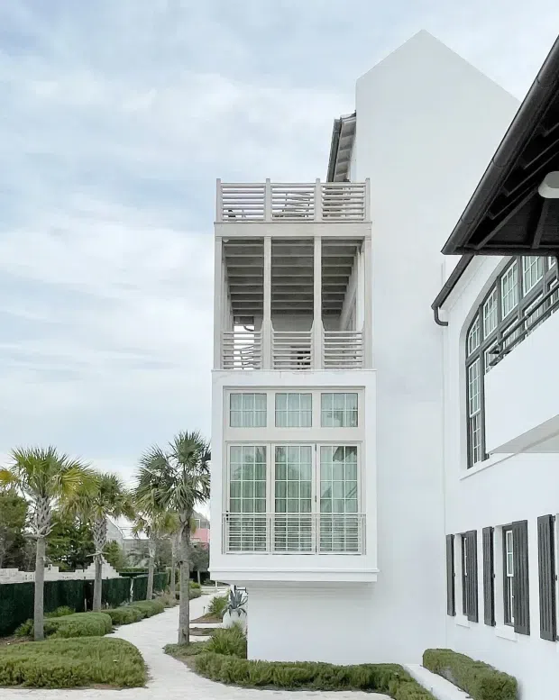 Exterior view of a tall, narrow white multi-storey building with stacked balconies, horizontal slatted railings, extensive gridded glazing, and dark window shutters. A landscaped pedestrian path with palm trees and clipped hedges runs alongside the building. The image emphasizes the bright coastal-style facade and vertical balcony tower.
