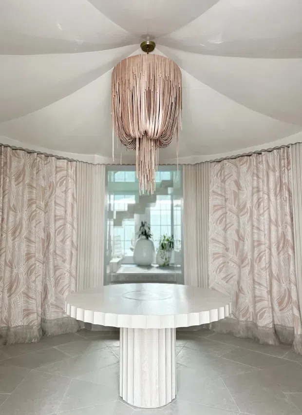 A bright, highly stylised round dining room with a large circular pedestal table, sculptural pleated edge, and a dramatic suspended looped-strand pendant light. The room has curved walls and a curved window bay, floor-to-ceiling patterned curtains in soft blush and cream, a white faceted ceiling, and large grey stone-look floor tiles. A reflective opening at the rear shows decorative vases and plants.