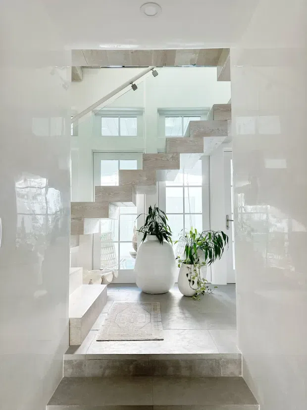 A bright, minimalist interior view through a wide opening toward a sculptural floating staircase. The space features pale stone or concrete-look tiled floors and stair treads, glossy white walls, large windows, and oversized white planters with indoor greenery. A low built-in seating or platform element is visible on the left, while the staircase spans diagonally across the double-height space.
