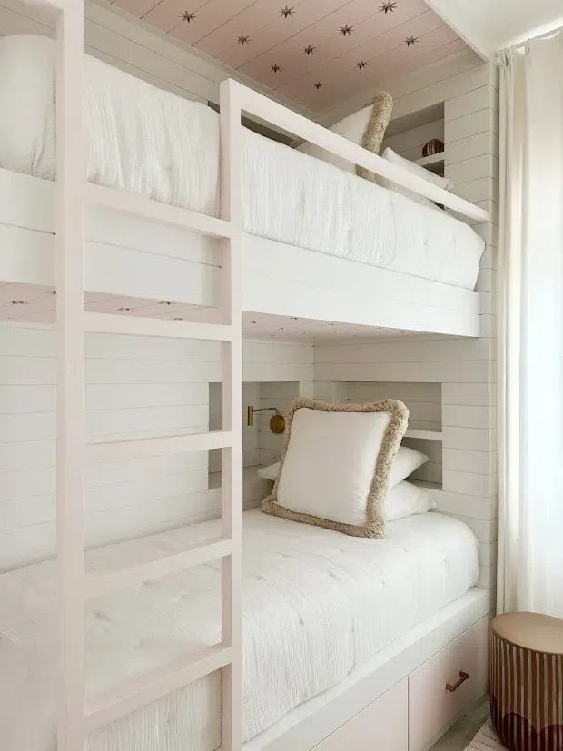 A compact bunk bedroom with custom built-in white bunk beds, ladder, and shiplap-style wall panelling. Both bunks are dressed in white textured bedding, with a plush-edged decorative pillow on the lower bunk. The ceiling has a pale pink finish with small star-shaped accents, while sheer curtains provide soft daylight at the window. The overall design is light, playful, and tailored for children.