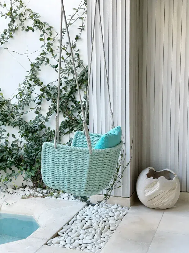 A tranquil outdoor poolside setting with a suspended woven hanging chair in pale aqua, styled with a matching cushion beside a white vertical-panel wall. Lush climbing greenery, white decorative pebbles, pale paving and a glimpse of blue pool water create a light, coastal-inspired atmosphere.