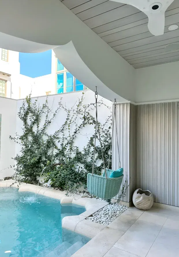 Bright contemporary covered outdoor pool courtyard with a curved white ceiling, pale vertically slatted wall, light tiled paving, pebble drainage bed, climbing greenery, and a turquoise hanging swing chair beside a small turquoise swimming pool. The space has a clean, sculptural, resort-like feel with strong indoor-outdoor connection.
