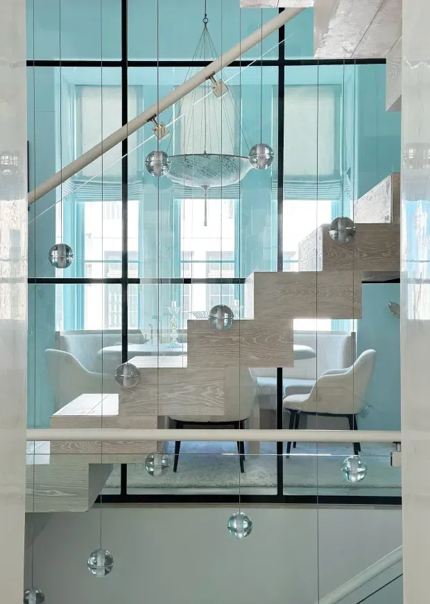 A bright, high-ceilinged contemporary interior viewed through a dramatic glazed stair enclosure. Pale timber stair treads and landings zigzag through the frame, with black-framed floor-to-ceiling glass panels, a pale blue and white palette, and multiple suspended spherical pendant lights. A dining area with a round table and upholstered chairs is visible behind the stair, creating a refined, airy architectural composition.