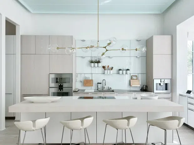 Bright, spacious contemporary kitchen with a large white island and four sculptural stools. Pale wood-grain cabinetry spans the back wall, with integrated ovens, refrigerator, sink, tiled splashback, open shelving, and a linear sculptural pendant light. White walls, a light-toned floor, and generous daylight create a clean, refined atmosphere.