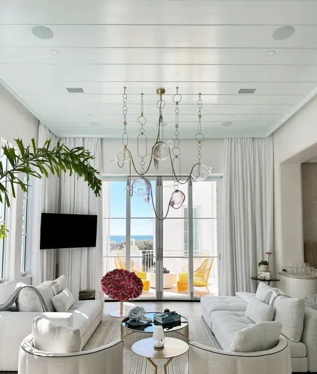 Bright, refined living room with floor-to-ceiling curtained glazing and French doors opening to a balcony with ocean views. The room features pale upholstered sofas and lounge chairs, a neutral rug, layered round coffee tables, sculptural hanging pendant lights, indoor greenery, and a wall-mounted television. A textured feature wall and drinks display are visible at right.
