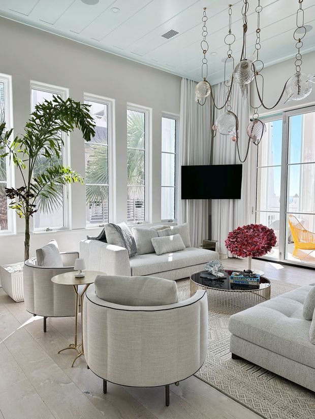 Bright, airy living room with tall multi-pane windows and glazed doors, pale upholstered seating, a patterned area rug, indoor palms, sheer curtains, a wall-mounted television, and a distinctive hanging sculptural glass-and-metal chandelier. The room has a refined coastal feel with abundant natural light and views of greenery and a balcony beyond the glazing.