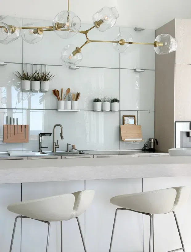 Bright contemporary kitchen with a long white island and two sculptural bar stools. The kitchen features glossy white full-height cabinetry and splashback panels, pale wood-toned cabinetry at the right, stainless-steel fixtures, open shelving with potted plants and utensils, and a distinctive branching brass pendant light with clear glass globes.