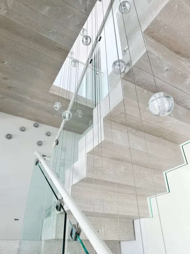 Interior architectural view looking upward through a tall stairwell or landing void. A sculptural staircase with glass balustrades and a white handrail is bordered by extensive floor-to-ceiling glazing, while multiple clear spherical pendant lights hang at different heights. The ceiling and soffits feature pale, textured timber-look panels, creating a bright, airy contemporary space.