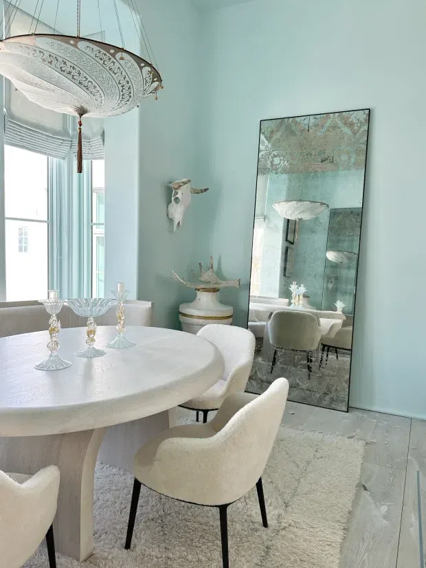 A bright, refined dining room in a pale blue and ivory palette. A round sculptural dining table is surrounded by upholstered boucle-style chairs, with crystal candleholders and a large ornate pendant light above. Floor-to-ceiling glazing with sheer Roman blinds brings in abundant daylight. A tall decorative mirror, sculptural wall objects, a console, textured rug, and pale timber flooring complete the room.