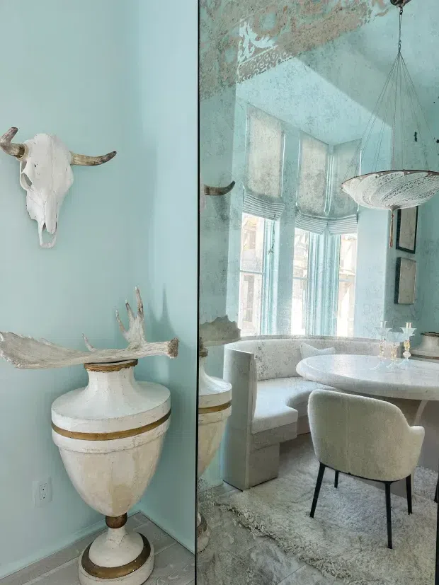 A refined, pale blue dining room vignette featuring a large mirrored wall, a round white dining table with upholstered curved chairs, a banquette, and an ornate suspended light fixture. The adjoining wall displays a decorative white animal skull and a sculptural antler arrangement on a pedestal urn, creating a dramatic eclectic focal point. Tall windows with pale patterned Roman blinds bring in soft daylight, while the mirror visually enlarges the room.