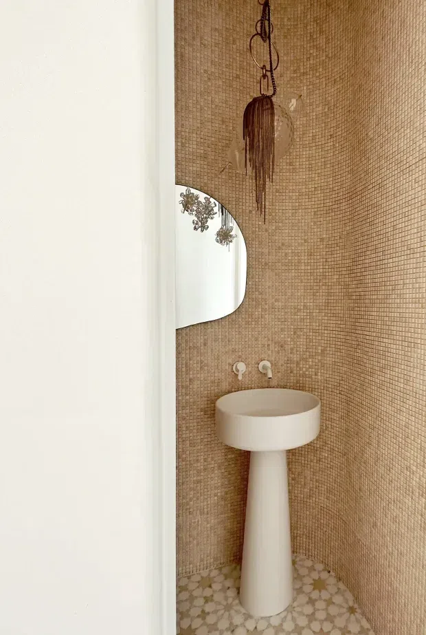 A compact powder room with a sculptural white pedestal basin and wall-mounted white taps. The walls are covered in warm tan mosaic tiles, while the floor has a patterned geometric tile finish. A rounded rectangular mirror and a decorative hanging light or ornament add visual interest. The space has a warm, refined, contemporary-organic feel.