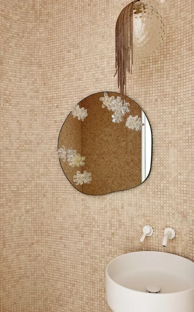 A close-up view of a refined bathroom vanity area. The walls are fully clad in small pale blush-beige mosaic tiles, with a rounded white vessel basin and matching wall-mounted tapware. A large organically shaped mirror with a warm timber-toned frame is mounted above the basin and decorated with translucent floral ornaments. A sculptural pendant with hanging strands adds a soft, decorative focal point.