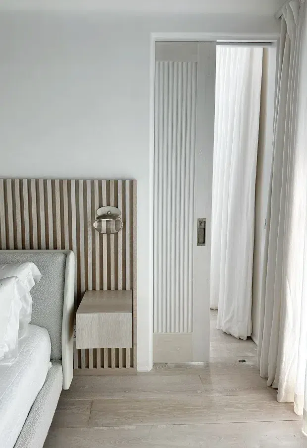 Bright, softly styled bedroom corner with a pale upholstered bed, white bedding, a vertically slatted timber feature headboard, and a matching light wood floating bedside table. Sheer white curtains cover tall glazing or a doorway, while a white ribbed sliding door and light timber-look flooring create a calm, tonal interior.