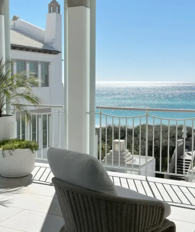 Bright coastal balcony or terrace overlooking the ocean, with white columns, balustrades, pale tiled flooring, and a woven lounge chair in the foreground. A white rendered building with a pitched roof and chimney is visible to the side, while landscaped rooftops and the sparkling turquoise sea extend beyond.
