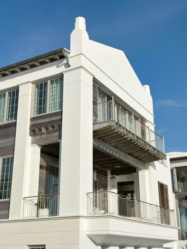 Exterior view of a tall white rendered building with strong vertical pilasters, stepped parapet elements, a prominent chimney-like feature, large multi-pane windows, timber-clad sections, and multiple projecting balconies with slender metal balustrades. The facade has a refined early-20th-century-inspired character, photographed in bright daylight against a blue sky.