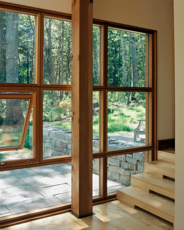 Interior view of a warm, wood-framed glazed space opening onto a wooded landscape and stone terrace. Floor-to-ceiling windows with timber mullions surround a substantial exposed timber post, while broad timber steps lead upward at right. Pale tiled flooring near the glazing transitions to light timber flooring by the steps. The space is bright, natural, and closely connected to the outdoors.