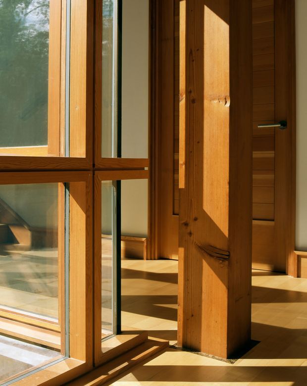 Sunlit interior architectural detail featuring substantial exposed timber posts, timber-framed glazing, a timber door with a modern metal lever handle, and warm timber flooring. Strong angular sunlight creates geometric shadows across the floor and walls. The image emphasizes the building fabric and circulation/opening rather than a furnished room.