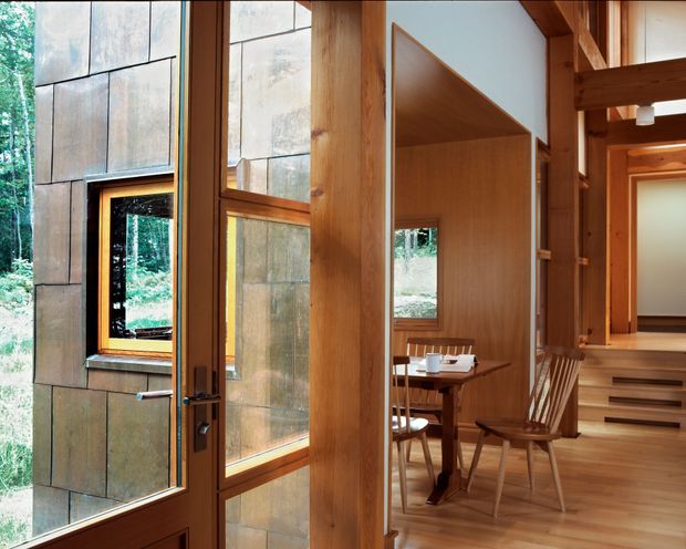 Interior view of a warm timber-framed home with a glazed entry door, large windows, exposed structural beams, and a compact dining area visible beyond. The space features extensive natural wood joinery and flooring, a stone-clad exterior wall visible through the glazing, and steps leading toward another interior area. The setting appears closely integrated with a wooded landscape.