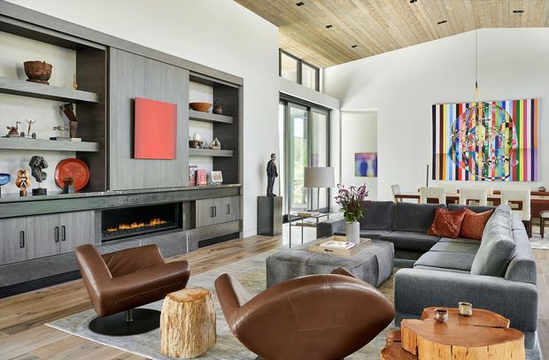 A spacious contemporary open-plan living room with a large grey sectional sofa, brown leather lounge chairs, timber stump-style side tables, and a central upholstered ottoman. A full-height built-in entertainment wall incorporates grey cabinetry, open display shelving, a long dark stone-like ledge, and a linear fireplace. Tall glazing brings in natural light, while a timber-lined ceiling, colourful abstract artwork, and a dining area beyond add visual interest.