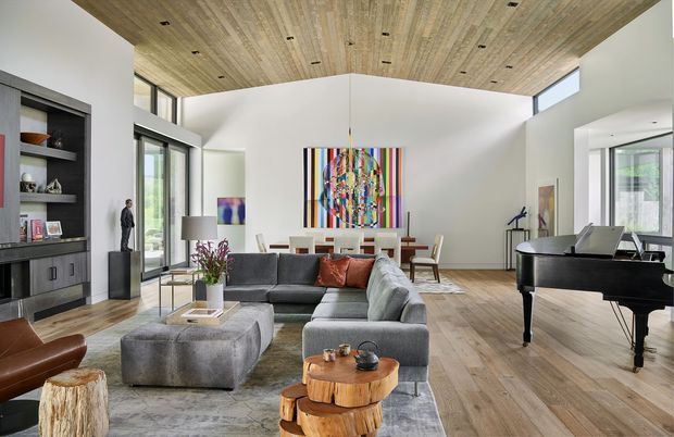 A spacious contemporary open-plan living room with a high angled timber-lined ceiling, white walls, extensive black-framed glazing, and wide-plank timber flooring. A large grey sectional sofa and ottoman face a low fireplace or media wall with built-in dark joinery and shelving. A grand piano occupies the right side, while a dining area and colourful abstract artwork sit at the rear. The room is bright, refined, and art-focused, with natural timber and neutral furnishings accented by vivid artwork and warm-toned leather seating.