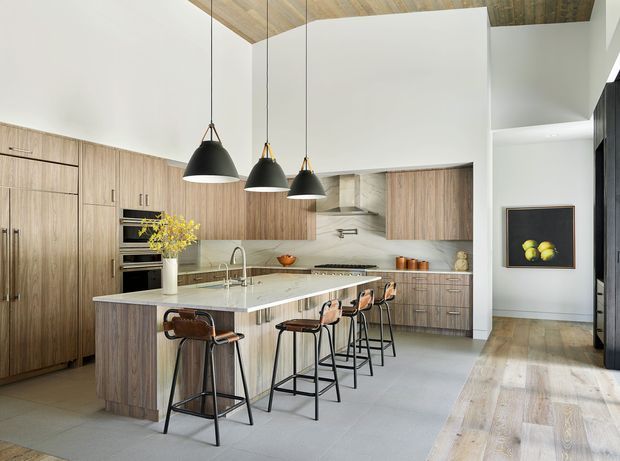 A spacious contemporary kitchen with a large wood-grain island, pale stone benchtop, integrated timber cabinetry, stainless steel appliances, and four bar stools. Three black pendant lights hang above the island. The kitchen opens toward a bright living or circulation area, with a timber-lined ceiling, pale tiled flooring, and timber flooring beyond.