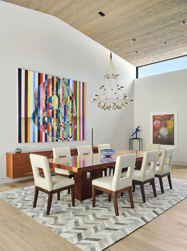 Bright, high-ceilinged dining room with a large rectangular dark timber dining table surrounded by upholstered cream dining chairs. A long mid-century-style timber sideboard sits against the white wall beneath a bold multicoloured abstract artwork. The room features a sculptural branching pendant chandelier, pale timber flooring, a geometric grey-and-cream rug, recessed ceiling lights, and floor-to-ceiling glazing at the upper right. The overall look is refined, colourful, and contemporary with mid-century influences.