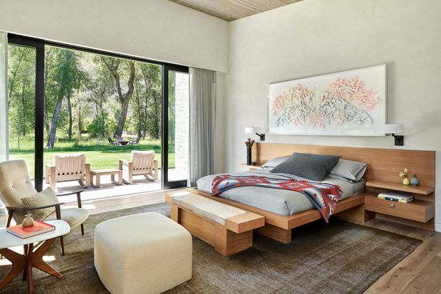 A spacious, softly lit bedroom with a low-profile timber platform bed and matching integrated bedside cabinetry. Large black-framed glazed doors open the room toward a leafy outdoor setting, while a neutral wool rug, upholstered lounge chair, ottoman, and abstract artwork create a calm contemporary retreat. The ceiling has a subtle linear timber finish and the room features warm timber flooring and joinery.