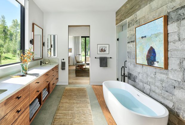 Bright, spacious contemporary bathroom with a freestanding white bathtub, long timber vanity with integrated basins, wall mirrors, and a walk-in shower area. A rugged grey stone feature wall with timber-toned upper cladding contrasts with white walls and warm timber flooring. Large glazing brings in abundant natural light and views of greenery, creating a refined yet organic atmosphere.