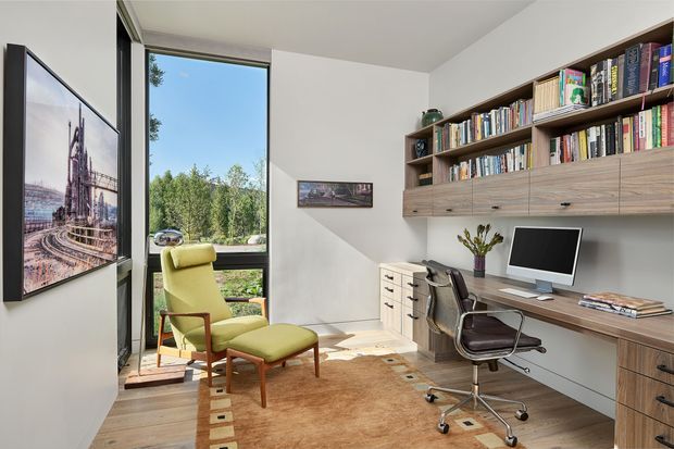 Bright, compact home office with a built-in timber desk and extensive wall-to-wall bookshelves, computer workstation, ergonomic office chair, and a vintage-style lounge chair with ottoman beside full-height glazing. The room has white walls, timber flooring, warm wood joinery, and a patterned area rug, with views to greenery outside.