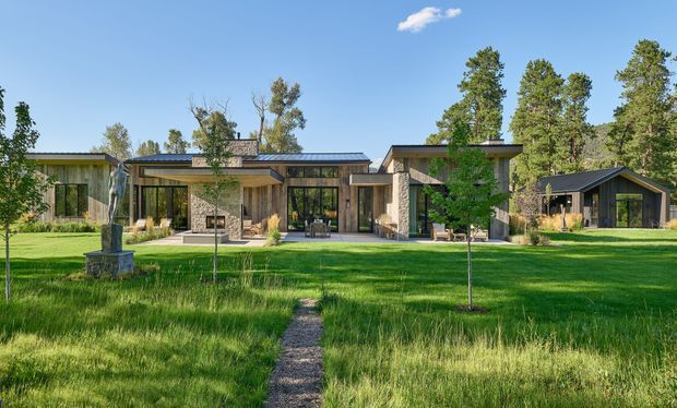 Wide exterior view of a low-slung contemporary residence set within a spacious landscaped lawn. The home has multiple connected wings, flat and shallow-pitched roof forms, extensive dark-framed glazing, natural stone or masonry feature walls, and warm timber cladding. A narrow gravel path leads through the grass toward a recessed central entry and covered outdoor living area. Mature trees and surrounding greenery frame the property.