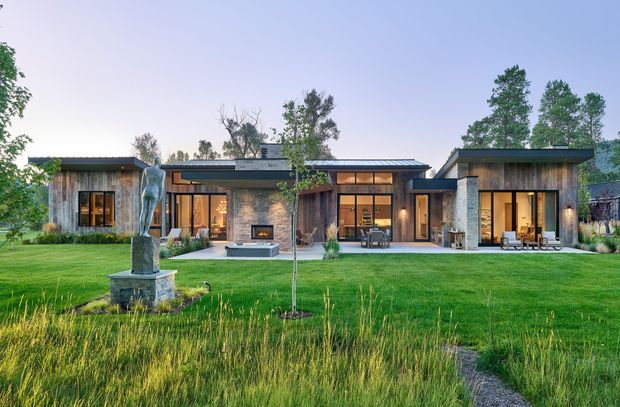 A wide exterior view of a low, single-storey contemporary residence set within a landscaped lawn. The home has a strongly horizontal composition with several connected wings, dark low-pitched roofs, extensive warm-lit glazing, natural stone walls, and timber-clad sections. A sculptural artwork stands in the foreground, with ornamental grasses, young trees, and a narrow garden path framing the house.