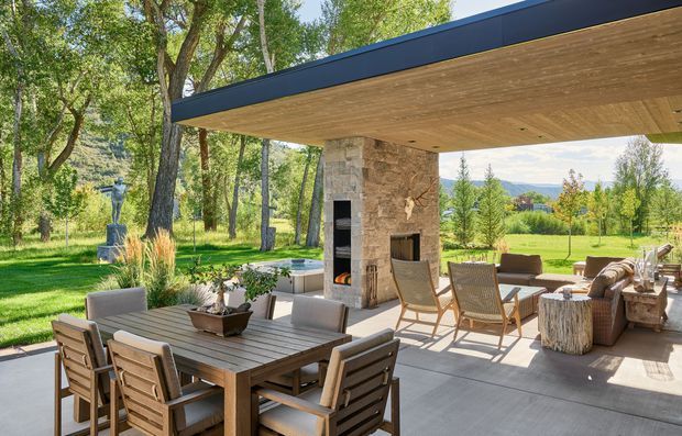 A spacious covered outdoor entertaining area beside a landscaped garden, featuring a flat roof with a warm timber soffit, dark fascia, stone-clad fireplace column, outdoor dining setting, lounge seating, and a built-in grill or fireplace opening. The contemporary pavilion opens broadly to mature trees, lawn, and distant mountain scenery.