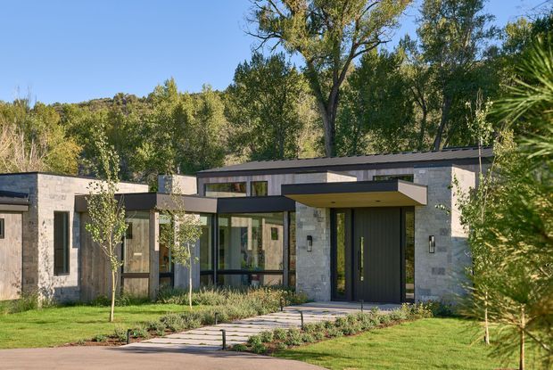 A low, modern single-storey residence set within a landscaped suburban or wooded site. The facade combines pale grey masonry with extensive dark-framed glazing, a broad flat roof, and projecting dark horizontal canopy forms over the entrance and living areas. A paved path leads through lawn and planting to a recessed front door, with mature trees and dense vegetation behind the house.