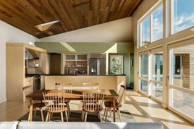 A bright, open-plan kitchen and dining area with a warm timber-clad ceiling, pale built-in cabinetry, dark benchtops, stainless-steel appliances, and a timber dining table with spindle-back chairs. Large floor-to-ceiling windows and glazed doors provide abundant natural light and views to an exterior deck. The room has a high, partially vaulted ceiling and a contemporary natural-material palette.