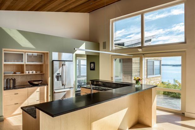 Sunlit contemporary kitchen with a large L-shaped island, pale wood cabinetry, dark stone benchtops, stainless-steel appliances, open shelving, and expansive windows and glazing overlooking a body of water. The room has a high slatted timber ceiling and a bright, airy connection to an exterior deck or terrace.